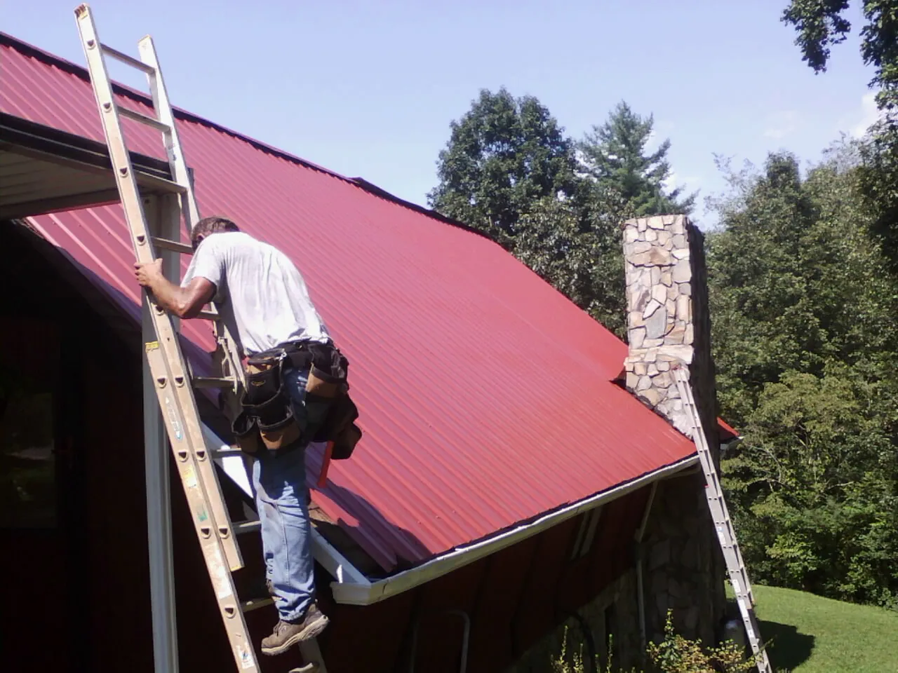 Roofer on a ladder beside a red metal roof and stone chimney — Soffit & Fascia Repair by Roofing Brothers in Goldston